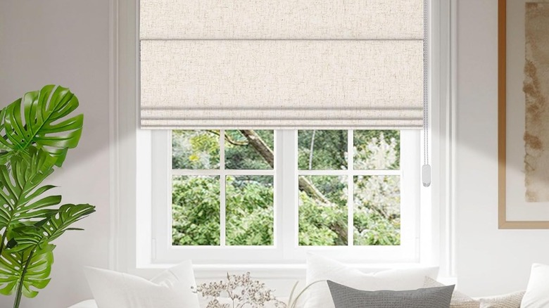 A white living room window is covered with off-white roman shades.