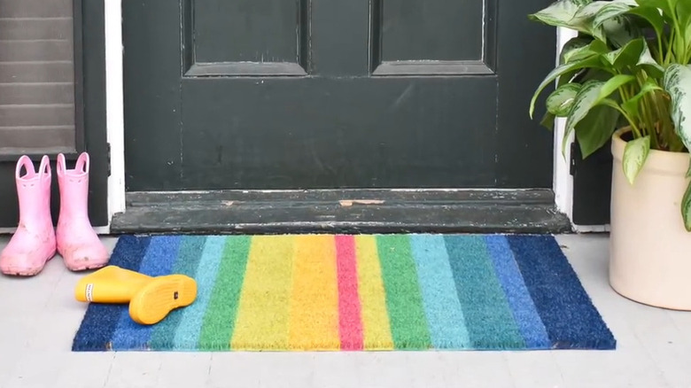 Rainbow painted doormat in front of a black door next to a pair of pink rain boots and a single yellow rainboot