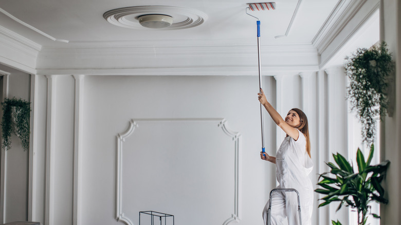 A woman dressed in white using a long paint roller to apply white paint to a ceiling.