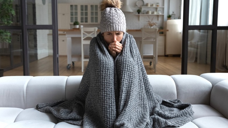 cold girl wearing gray winter hat while wrapped up in blanket on living room sofa