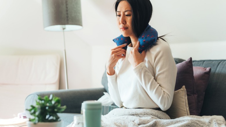 Woman sitting on couch with blanket over lap and heating pack on neck