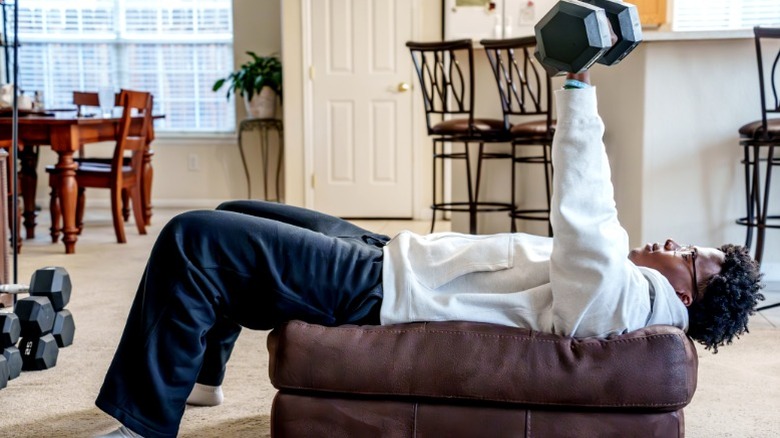 Man in hoodie and sweats doing presses with free weights on living room ottoman