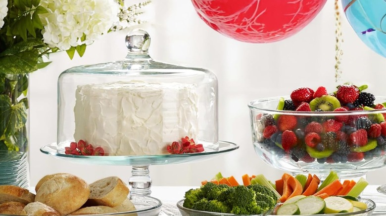 A domed cake stand is surrounded by party food and balloons