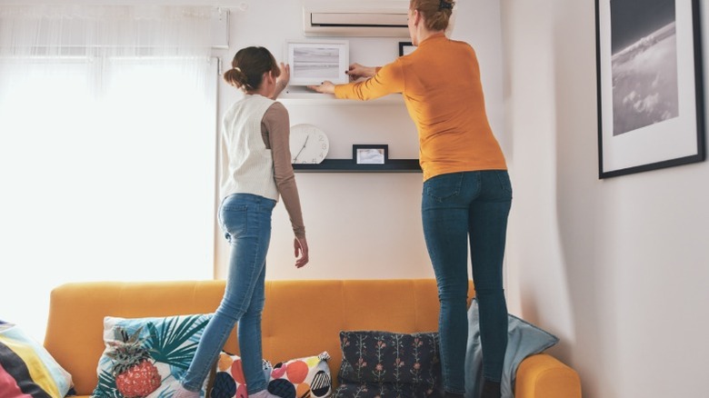 Two people standing on a yellow couch decorating shelfs above it with frames.