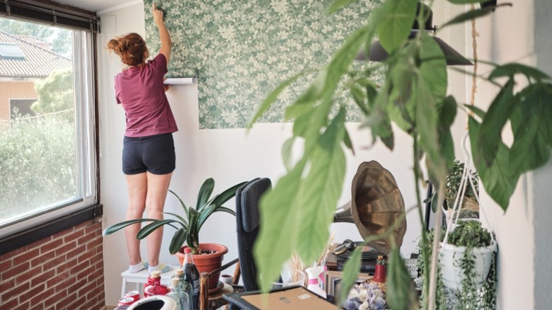 A woman sticking green floral wallpaper onto a wall in a room with plants and other items.