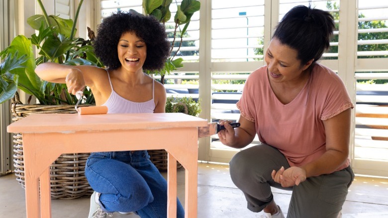 Two people painting a small table a peach color with a large houseplant behind them.