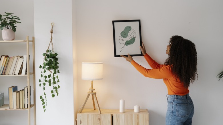 A woman hanging up an art print of an abstract leaf over a short cabinet.