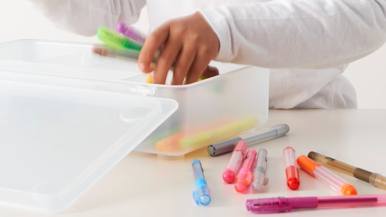 Plastic box with an attached lid with hands putting pens inside