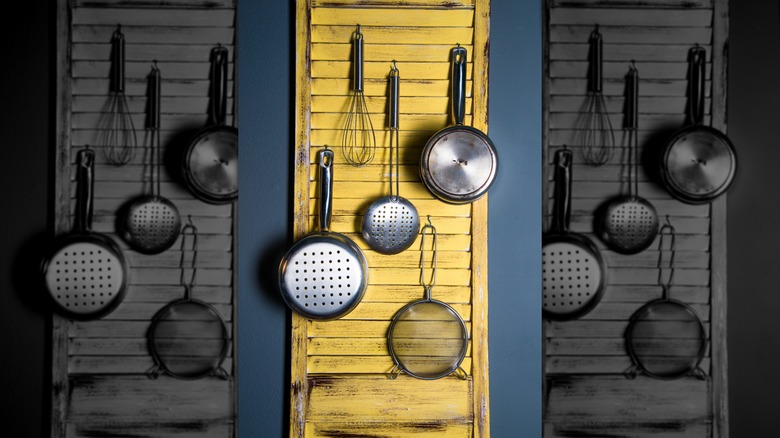 An old yellow shutter has various kitchen pots and tools hanging on it from hooks.