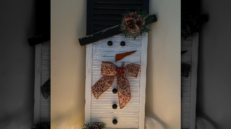 An adorable snowman made from a simple shutter is donned with a holiday ribbon bow and snow dusted garland accents.