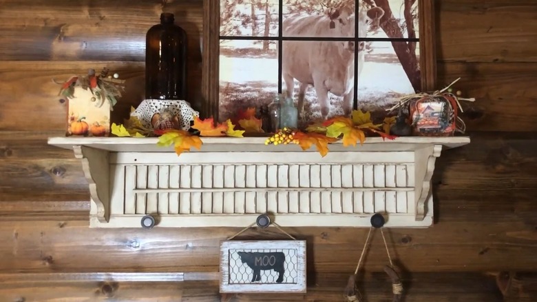 A distressed white shutter is attached to the back of a L-mounted shelf with fall decorations on top.