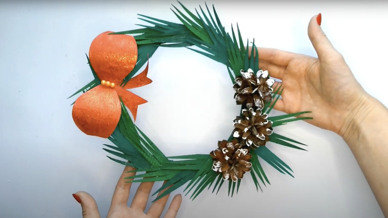 A person holds a wreath made of cardboard spikes with a red sparkly bow and pinecones.