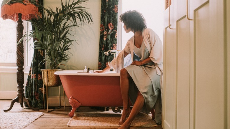 A woman gets ready for a bath in a luxurious bathroom with an orange-colored tub and vintage accents