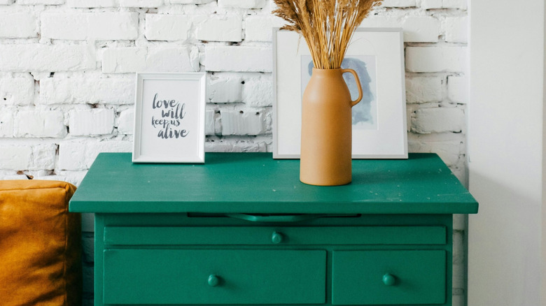 Some decor sitting on a green-colored nightstand in front of a white brick wall.