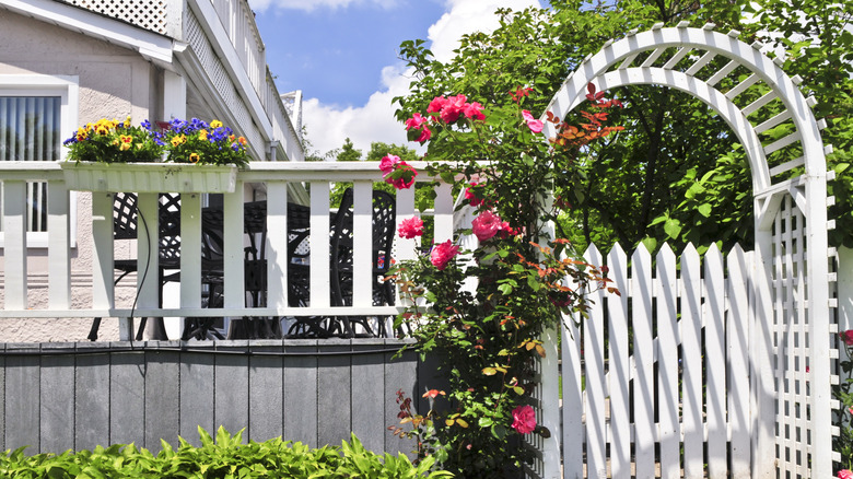 Pretty white fence gate with an arbor next to the deck of a house surrounded by colorful flowers