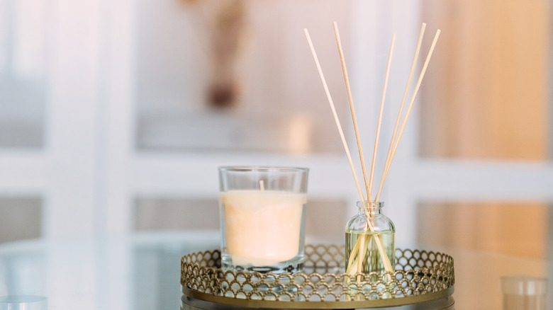 A reed diffuser in a small perfume glass bottle sits on a table next to a candle