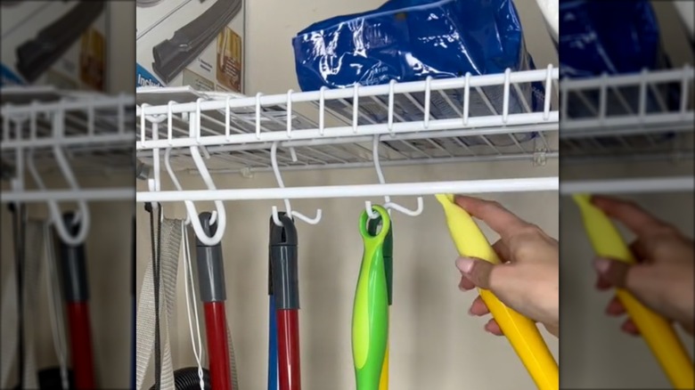 White storage rack in a closet that has the handles of brooms and other cleaning items hanging from S-hooks