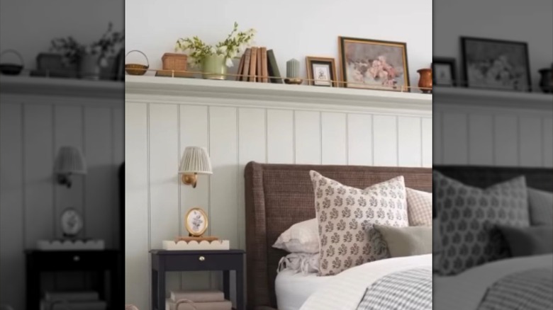 A white wainscoting wall behind a bed with a display shelf on the top featuring a decorative rail.