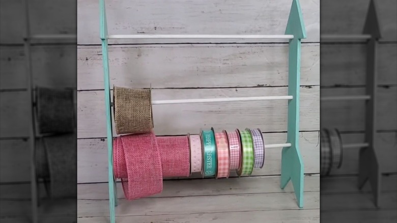 A DIY craft robbon organizer with light blue sides in the shape of arrows and three wooden dowels across them holding colorful ribbon.