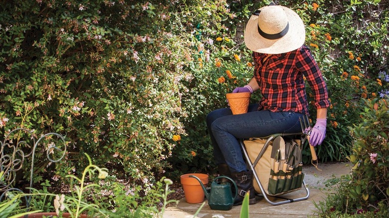 A person sits on a folding chair that doubles as bag holding a set of garden tools.