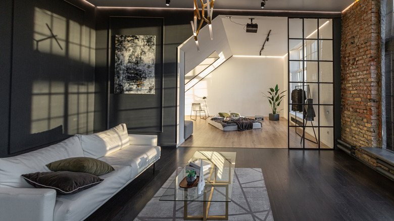 A darkly gray painted living room with a brick wall, white furniture and brown accents with sunlight coming in the window.