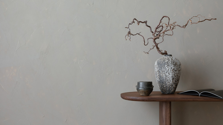 A wooden side table with an artisanal wooden plant and stone vase sitting in front of a light gray wall.