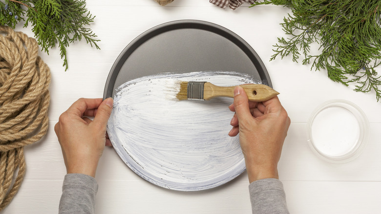 Hands are prepping an aluminum pan with white paint before adding greenery, rope, and a bow for a festive DIY wreath.