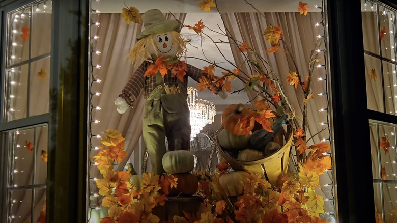 A bay window filled with fall decor including fall leaves, a scarecrow, pumpkins, and lights