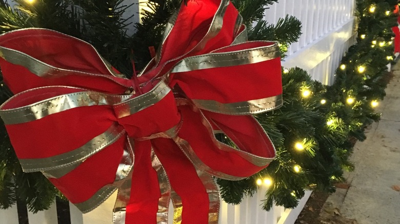 Large red and gold bow on corner of white picket fence with lighted garland