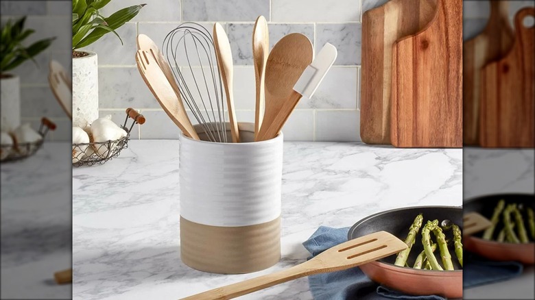 A white and tan ceramic crock holds wooden utensils in a marble kitchen