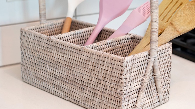 Pink and wooden utensils are stored in a rattan caddy on a countertop