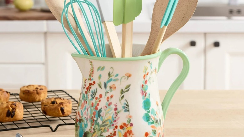 A floral pitcher holding kitchen utensils sits next to muffins on a cooling rack