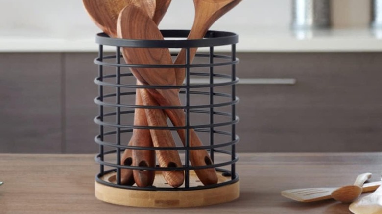 A bowl of fruit sits next to a wire utensil holder on a wooden countertop