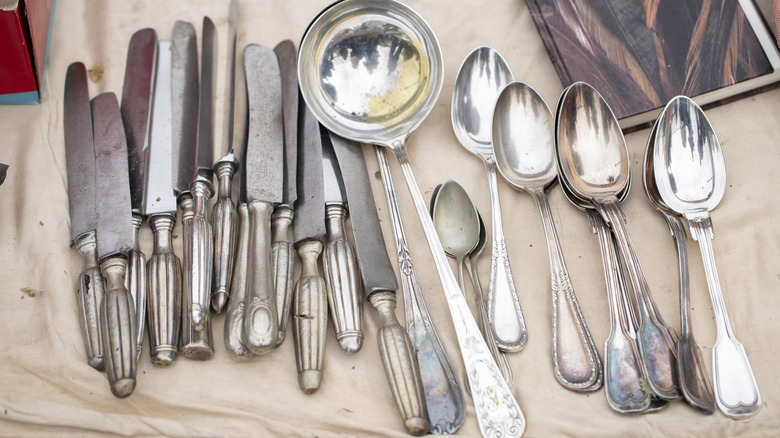 A set of tarnishing silver butter knives, a ladel, and spoons laying on a canvas tarp.