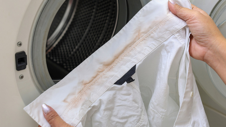 Hands holding a white blouse with discoloration at the collar in front of a washing machine.