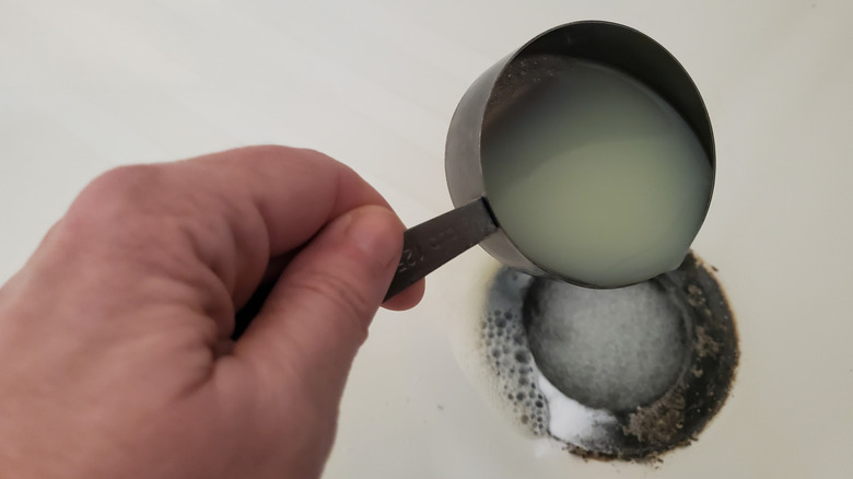 A Caucasian hand pours lemon juice into a shower drain already filled with baking soda.