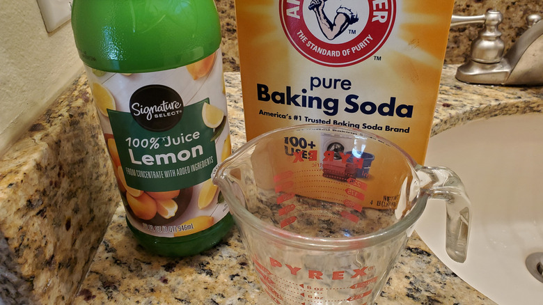 A bottle of lemon juice, a box of baking soda, and a measuring cup sit on a bathroom counter.