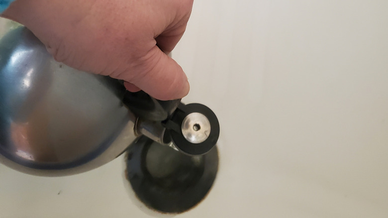 A Caucasian hand holding a kettle pours boiling water into a shower drain.