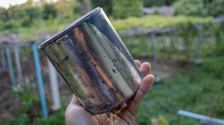 A person holds a metal container while standing in the garden.