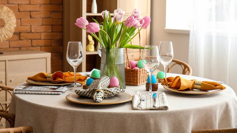 A round dining table set with gold napkins, colorful egg decor, and a vase of pink tulips