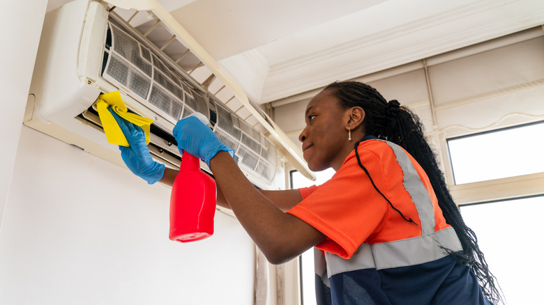 Woman spraying air conditioner with cleaner and wiping with cloth