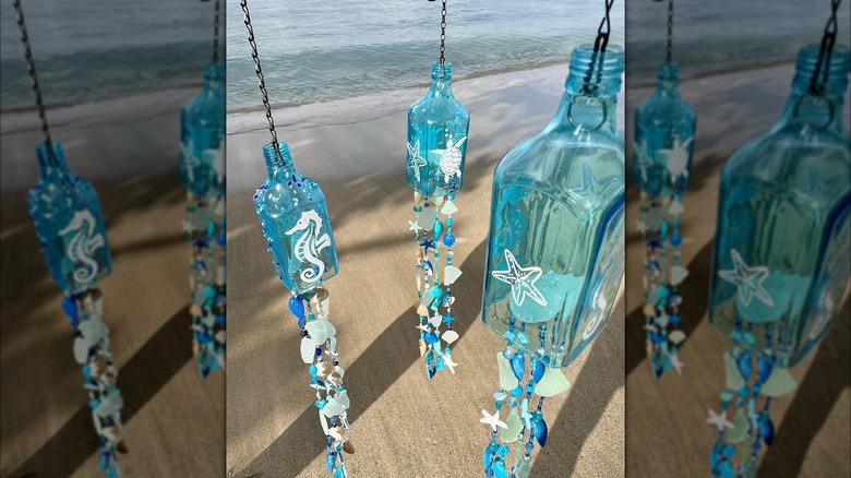 Teal glass bottles cut and turned into ocean-themed wind chimes being held in front of the ocean.