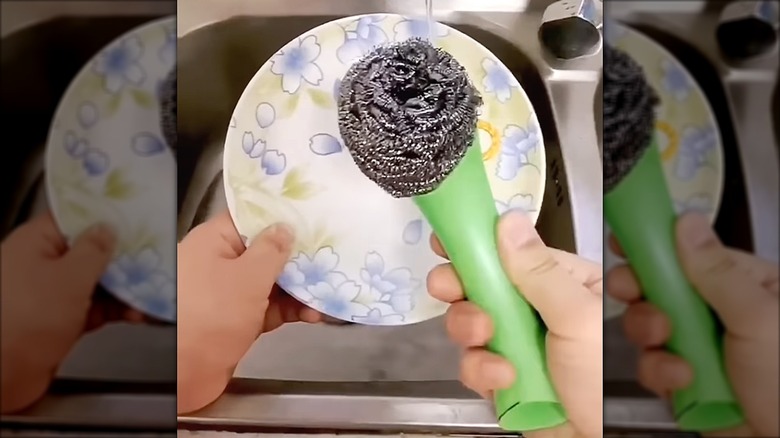 Person holding and washing dishes with a scrub brush made from the handle of a plastic vinegar jug.