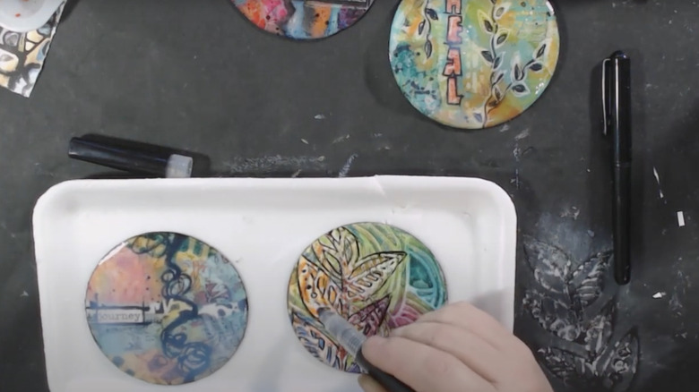 A person draws on old coasters to create decorative fridge magnets.