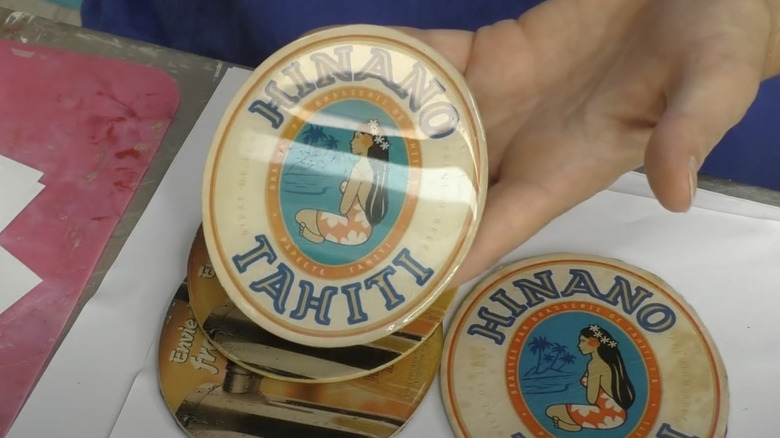 A hand holds up an oval cardboard bar coaster with a shiny resin surface.