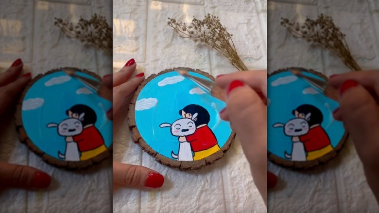 A person paints a cartoon on a wooden coaster.