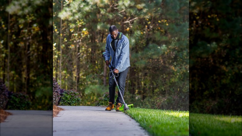 A man uses a Greenworks string trimmer on grass along the edge of a concrete walkway