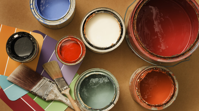 Variety of different colors and types of paints in open cans, next to paint brushes and color swatches