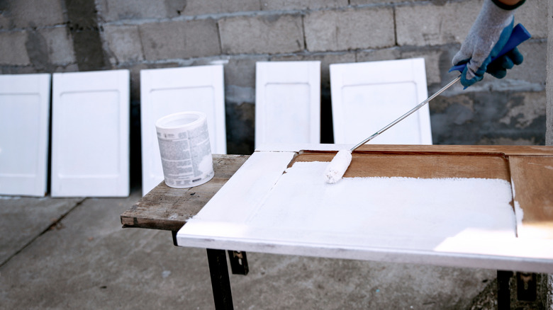 Painted cabinets drying in background while gloved hand paints cabinet door with roller