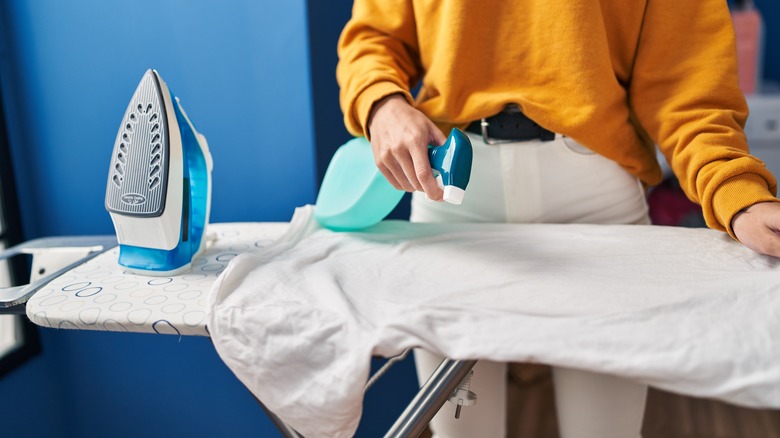 woman spraying white shirt with blue spray bottle on ironing board